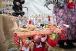 A table is adorned with colorful handmade dolls and crafts, including a zebra mobile and eclectic fabric items. The dolls are dressed in vibrant traditional attire, featuring reds, blues, and whites. Surrounding the dolls are small fabric balls and decorative items. The setup suggests a market or a craft fair, with a background of people and more textiles.