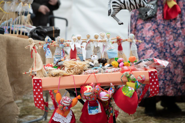 A table is adorned with colorful handmade dolls and crafts, including a zebra mobile and eclectic fabric items. The dolls are dressed in vibrant traditional attire, featuring reds, blues, and whites. Surrounding the dolls are small fabric balls and decorative items. The setup suggests a market or a craft fair, with a background of people and more textiles.