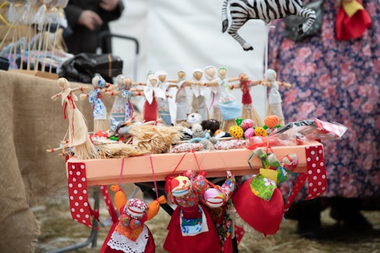 A table is adorned with colorful handmade dolls and crafts, including a zebra mobile and eclectic fabric items. The dolls are dressed in vibrant traditional attire, featuring reds, blues, and whites. Surrounding the dolls are small fabric balls and decorative items. The setup suggests a market or a craft fair, with a background of people and more textiles.