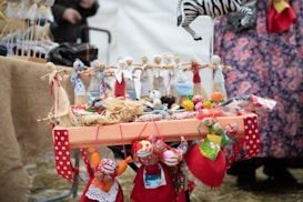 A table is adorned with colorful handmade dolls and crafts, including a zebra mobile and eclectic fabric items. The dolls are dressed in vibrant traditional attire, featuring reds, blues, and whites. Surrounding the dolls are small fabric balls and decorative items. The setup suggests a market or a craft fair, with a background of people and more textiles.
