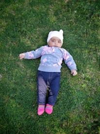 A baby lies on green grass, wearing a white hat, a gray sweatshirt with pink floral patterns, dark blue jeans, and bright pink socks.