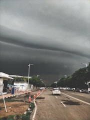 Dark, stormy clouds hang ominously over a straight urban road lined with traffic cones and cars. Trees border the sidewalk, and a few people can be seen near a white building with blue accents. The atmosphere is tense and anticipative of approaching rain.