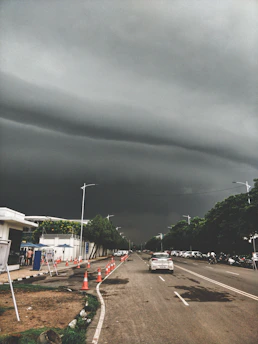 Dark, stormy clouds hang ominously over a straight urban road lined with traffic cones and cars. Trees border the sidewalk, and a few people can be seen near a white building with blue accents. The atmosphere is tense and anticipative of approaching rain.