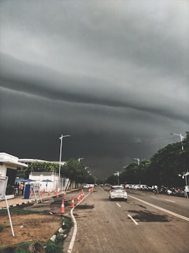 Dark, stormy clouds hang ominously over a straight urban road lined with traffic cones and cars. Trees border the sidewalk, and a few people can be seen near a white building with blue accents. The atmosphere is tense and anticipative of approaching rain.