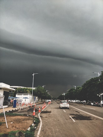 Dark, stormy clouds hang ominously over a straight urban road lined with traffic cones and cars. Trees border the sidewalk, and a few people can be seen near a white building with blue accents. The atmosphere is tense and anticipative of approaching rain.