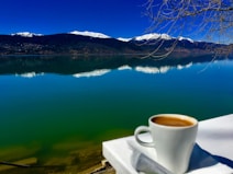 A serene lakeside view with calm water reflecting the clear blue sky and snow-capped mountains in the background. A white cup filled with coffee is placed on a white surface in the foreground, adding a cozy touch to the natural scenery. Bare tree branches extend from the right side.