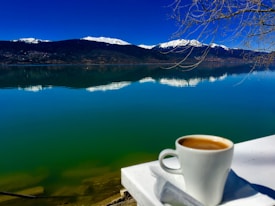 A serene lakeside view with calm water reflecting the clear blue sky and snow-capped mountains in the background. A white cup filled with coffee is placed on a white surface in the foreground, adding a cozy touch to the natural scenery. Bare tree branches extend from the right side.