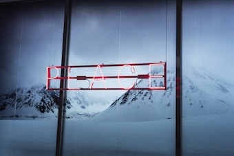 A neon sign displaying the word 'PEACE' is set against a serene backdrop of snow-covered mountains. The red glow of the sign contrasts with the muted blues and grays of the snowy landscape.