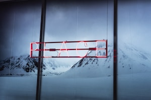 A neon sign displaying the word 'PEACE' is set against a serene backdrop of snow-covered mountains. The red glow of the sign contrasts with the muted blues and grays of the snowy landscape.