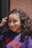 A person with curly hair smiles warmly while wearing a purple sweater with the word 'AMOUR' in bright red letters. The background features a brick wall and a dark door frame, providing contrast to the person's lively expression.