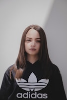 A young woman with long, straight brown hair stands against a plain white background. She is wearing a black Adidas sweatshirt with the brand's logo prominently displayed. The lighting creates a soft shadow on the wall, lending a contemporary and minimalistic aesthetic.