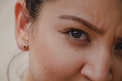 An extreme close-up of a person's face focusing on the eye and eyebrow area. The eye is expressive, with the eyebrow slightly furrowed, suggesting a thoughtful or concerned expression. A small stud earring is visible in the ear, and the skin has a natural, smooth texture.