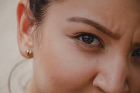 An extreme close-up of a person's face focusing on the eye and eyebrow area. The eye is expressive, with the eyebrow slightly furrowed, suggesting a thoughtful or concerned expression. A small stud earring is visible in the ear, and the skin has a natural, smooth texture.