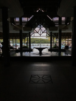 A shaded open-air structure with a high roof overlooking an outdoor pool area. Tropical greenery is visible in the background beyond the pool, creating a serene setting. The architecture is supported by columns, and a pond or water feature is centrally placed.