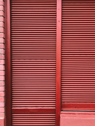Red metal shutters with horizontal ridges are framed by red and pink brick walls.