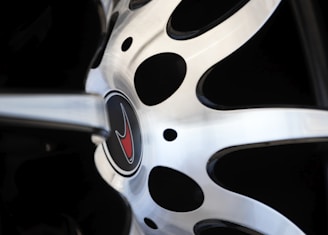 A close-up view of a car wheel rim with a metallic, polished surface. The center of the wheel features an emblem with a red logo, surrounded by a circular design with black and silver elements.