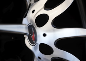 A close-up view of a car wheel rim with a metallic, polished surface. The center of the wheel features an emblem with a red logo, surrounded by a circular design with black and silver elements.