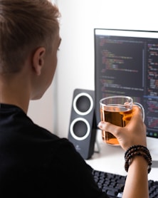 man holding clear glass mug while facing flat screen monitor
