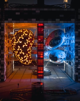 A modern architectural structure featuring two large, illuminated sculptures within a building. One sculpture is orange and resembles a cocoon or an intricate nest, while the other has a blue hue and appears to be an abstract, futuristic form. The space is illuminated with warm orange and cool blue lighting, creating a stark contrast. A red, vertical structure divides the two sculptures, adding to the modern aesthetic. A small, silhouetted figure is visible below, indicating the scale of the installation.