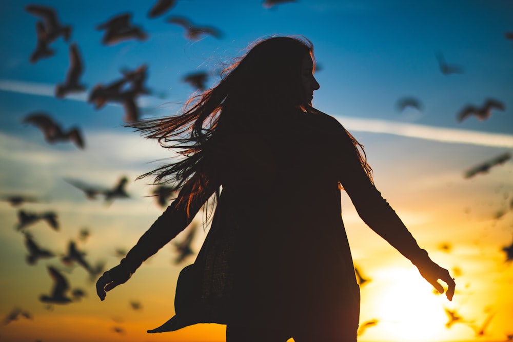 Silhouettenfotografie einer Frau, die ihre Hände vor dem Flug der Vögel ausbreitet