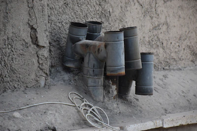 Close-up of rusted pipes intertwined with wires, dripping with dark liquid against a concrete wall.