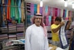 A man wearing a traditional white robe and red-checkered headscarf stands in front of stacks of colorful textiles displayed on shelves in a shop. Another person in a yellow shirt is folding or handling a piece of cloth next to him. Various vibrant fabrics in multiple patterns and colors are neatly arranged on the walls, creating a lively and colorful backdrop.