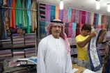 A man wearing a traditional white robe and red-checkered headscarf stands in front of stacks of colorful textiles displayed on shelves in a shop. Another person in a yellow shirt is folding or handling a piece of cloth next to him. Various vibrant fabrics in multiple patterns and colors are neatly arranged on the walls, creating a lively and colorful backdrop.