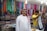 A man wearing a traditional white robe and red-checkered headscarf stands in front of stacks of colorful textiles displayed on shelves in a shop. Another person in a yellow shirt is folding or handling a piece of cloth next to him. Various vibrant fabrics in multiple patterns and colors are neatly arranged on the walls, creating a lively and colorful backdrop.