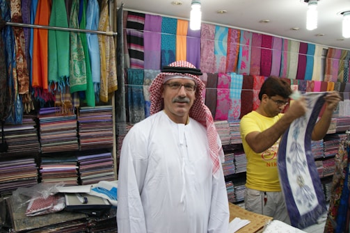 A man wearing a traditional white robe and red-checkered headscarf stands in front of stacks of colorful textiles displayed on shelves in a shop. Another person in a yellow shirt is folding or handling a piece of cloth next to him. Various vibrant fabrics in multiple patterns and colors are neatly arranged on the walls, creating a lively and colorful backdrop.