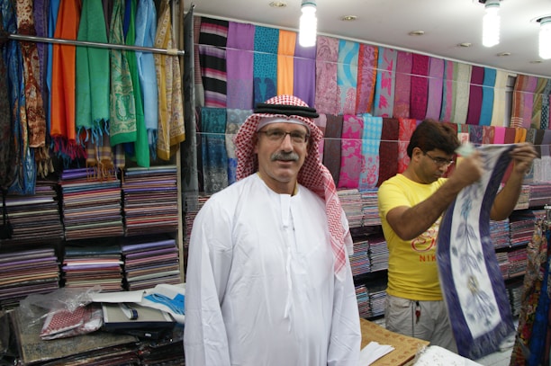 A man wearing a traditional white robe and red-checkered headscarf stands in front of stacks of colorful textiles displayed on shelves in a shop. Another person in a yellow shirt is folding or handling a piece of cloth next to him. Various vibrant fabrics in multiple patterns and colors are neatly arranged on the walls, creating a lively and colorful backdrop.