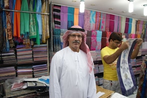 A man wearing a traditional white robe and red-checkered headscarf stands in front of stacks of colorful textiles displayed on shelves in a shop. Another person in a yellow shirt is folding or handling a piece of cloth next to him. Various vibrant fabrics in multiple patterns and colors are neatly arranged on the walls, creating a lively and colorful backdrop.