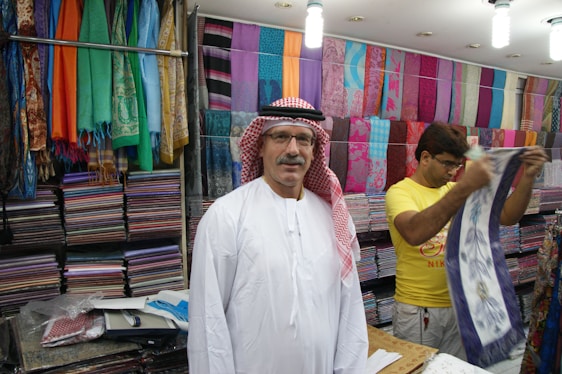 A man wearing a traditional white robe and red-checkered headscarf stands in front of stacks of colorful textiles displayed on shelves in a shop. Another person in a yellow shirt is folding or handling a piece of cloth next to him. Various vibrant fabrics in multiple patterns and colors are neatly arranged on the walls, creating a lively and colorful backdrop.