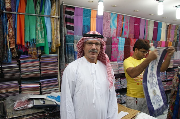 A man wearing a traditional white robe and red-checkered headscarf stands in front of stacks of colorful textiles displayed on shelves in a shop. Another person in a yellow shirt is folding or handling a piece of cloth next to him. Various vibrant fabrics in multiple patterns and colors are neatly arranged on the walls, creating a lively and colorful backdrop.