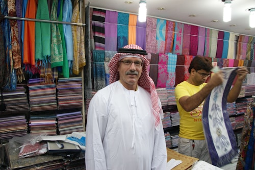 A man wearing a traditional white robe and red-checkered headscarf stands in front of stacks of colorful textiles displayed on shelves in a shop. Another person in a yellow shirt is folding or handling a piece of cloth next to him. Various vibrant fabrics in multiple patterns and colors are neatly arranged on the walls, creating a lively and colorful backdrop.