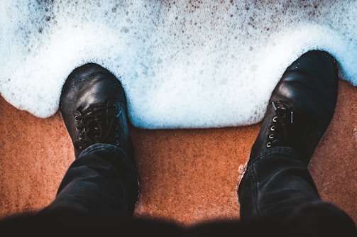 A pair of black leather shoes partially submerged in foamy seawater on a sandy beach. The image captures the moment when ocean waves gently touch the footwear, creating a contrast between the dark shoes and the white foam.