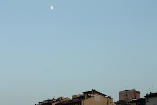 A row of buildings with varied roofs, antennas, and chimneys under a clear sky with a visible crescent moon.