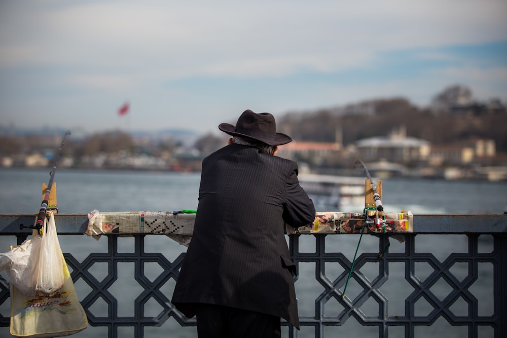 man leaning on handrail facing the body of water and buildings during day