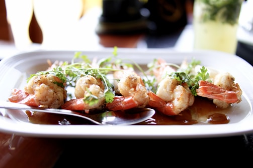 A plate of cooked shrimp is garnished with herbs and served with a dark sauce. The shrimp are positioned neatly on a rectangular white dish, with a spoon resting beside them. In the background, there is a blurred view of a refreshing glass with lemon and mint.