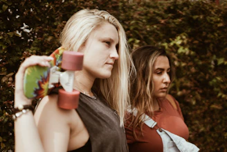 Two young women are standing outdoors against a leafy background. One woman is holding a skateboard over her shoulder and looking ahead solemnly, while the other woman stands beside her, looking down pensively. They both have casual attire and appear contemplative.