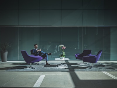 A man sits in a modern, minimalist office lobby. He is dressed in a suit and is looking at his phone. The lobby features two purple chairs and a small white table. A vase with colorful flowers is placed on the table. The ambiance is filled with natural light creating a contrast of shadows on the floor.