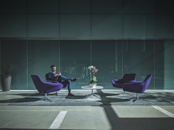 A man sits in a modern, minimalist office lobby. He is dressed in a suit and is looking at his phone. The lobby features two purple chairs and a small white table. A vase with colorful flowers is placed on the table. The ambiance is filled with natural light creating a contrast of shadows on the floor.