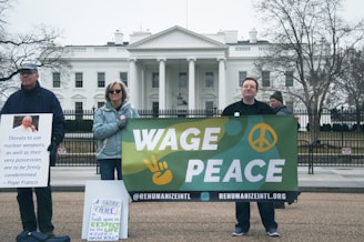 A group of people are standing in front of the White House holding signs. One person holds a sign with a quote from Pope Francis condemning nuclear weapons. Another individual holds a banner that reads 'Wage Peace' with a peace sign and a hand making a peace gesture. Another sign emphasizes respect for life. The atmosphere is calm and the trees are bare, suggesting a cold or winter setting.