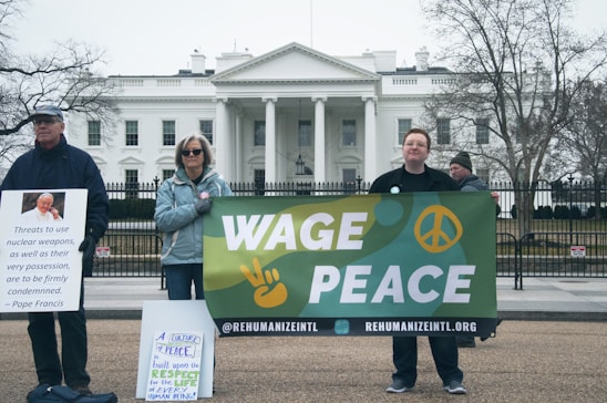A group of people are standing in front of the White House holding signs. One person holds a sign with a quote from Pope Francis condemning nuclear weapons. Another individual holds a banner that reads 'Wage Peace' with a peace sign and a hand making a peace gesture. Another sign emphasizes respect for life. The atmosphere is calm and the trees are bare, suggesting a cold or winter setting.