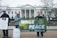 A group of people are standing in front of the White House holding signs. One person holds a sign with a quote from Pope Francis condemning nuclear weapons. Another individual holds a banner that reads 'Wage Peace' with a peace sign and a hand making a peace gesture. Another sign emphasizes respect for life. The atmosphere is calm and the trees are bare, suggesting a cold or winter setting.