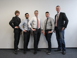 Five men are standing in a row indoors, all wearing formal attire including shirts, ties, and blazers. They are posing confidently with their hands on their hips against a plain white background. The setting appears to be an office space with a carpeted floor.