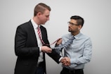 Two men are interacting in a light-hearted manner, with one man adjusting or holding the other's tie during a handshake. The setting appears casual yet professional, and both are wearing business attire.