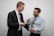 Two men are interacting in a light-hearted manner, with one man adjusting or holding the other's tie during a handshake. The setting appears casual yet professional, and both are wearing business attire.