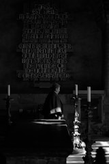 A person stands in what appears to be a dimly lit church or chapel. The surrounding area contains tall candlesticks and an altar. There is a large decorative text on the wall in the background, displaying a religious message in Latin and English. Light streams in, casting dramatic shadows and highlighting certain areas.