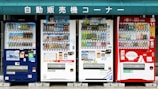 A row of four vending machines is displayed, each offering a variety of beverages. The machines are colorful, with numerous bottles and cans visible through the glass. Above the machines, there is a green sign with white Japanese text.