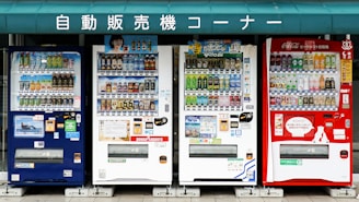 A row of four vending machines is displayed, each offering a variety of beverages. The machines are colorful, with numerous bottles and cans visible through the glass. Above the machines, there is a green sign with white Japanese text.